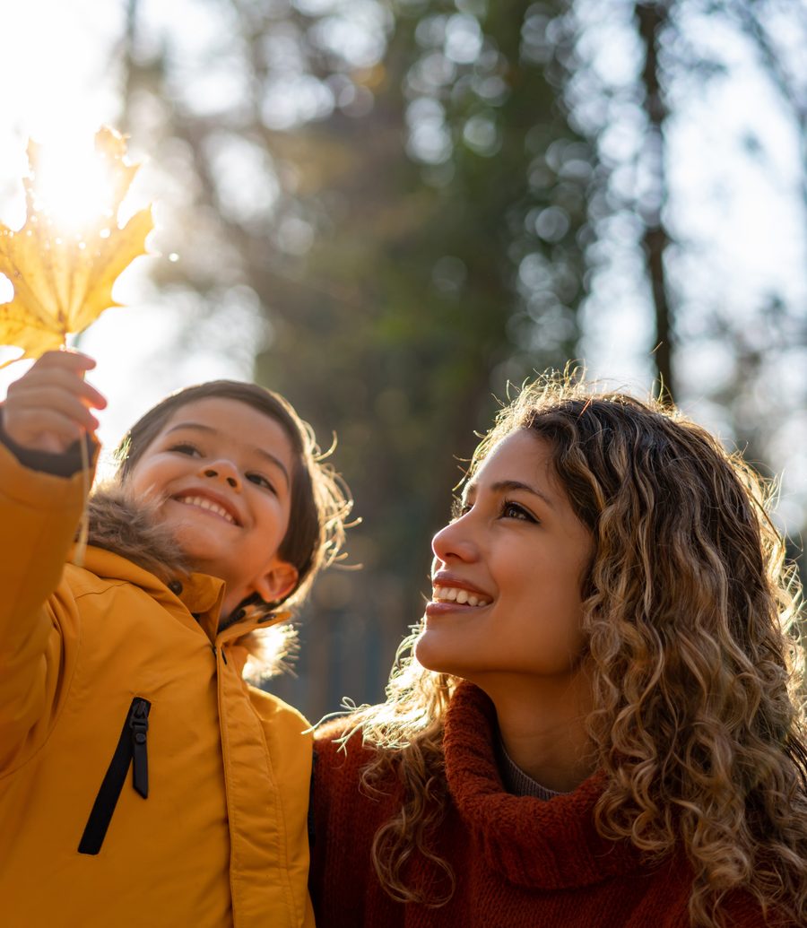 A mother and her young son smiling together outdoors on a sunny autumn day as he holds up a yellow leaf